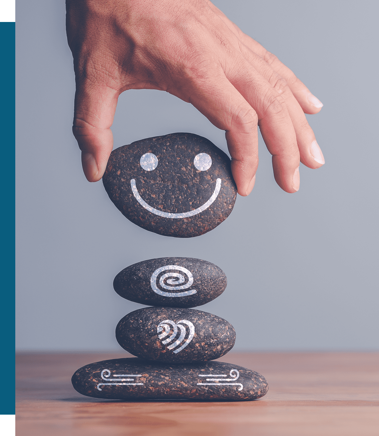 A hand places a stone with a smiley face drawn on it atop a stack of three decorated stones on a wooden surface.