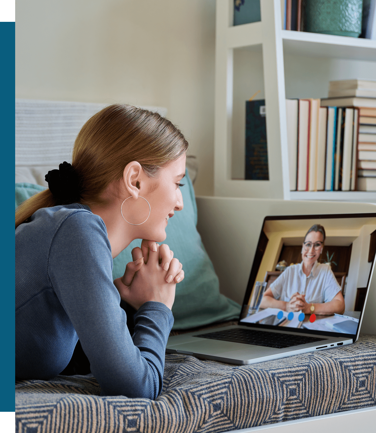 A woman lying on a bed smiles while having a video call with another woman, seen on the laptop screen in a home setting.