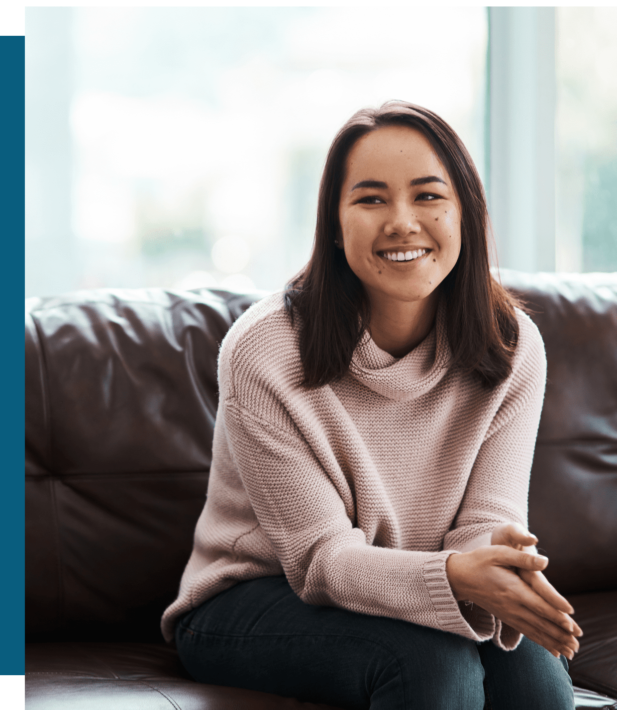 A woman with straight dark hair, wearing a light-colored sweater and jeans, sits on a brown leather couch and smiles at the camera.