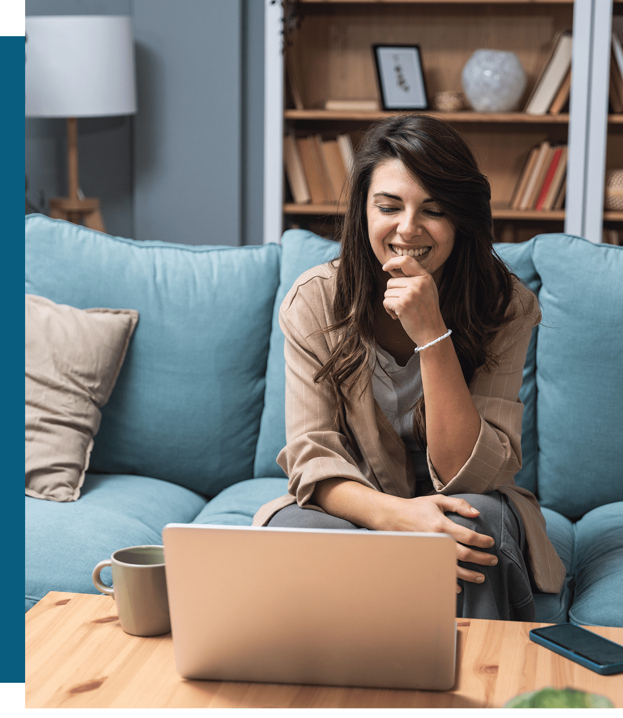 A woman sits on a blue sofa, smiling and looking at a laptop on a wooden table with a mug and smartphone nearby. A bookshelf is in the background.