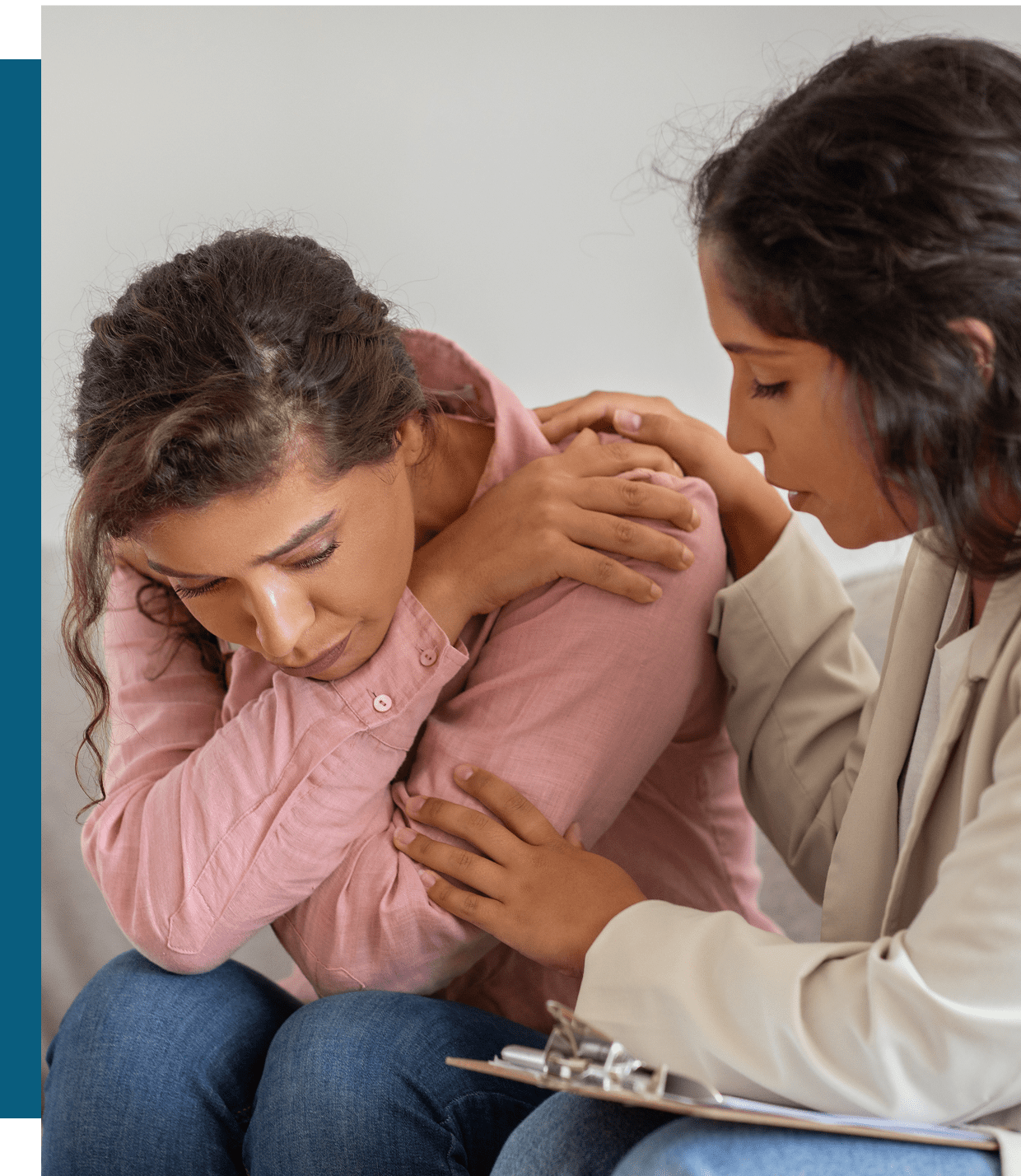 A woman sits with her head down and arms crossed, while another woman offers comfort by gently touching her arm and shoulder.